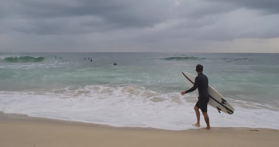 A surfer carries his board along a beach while watching the choppy waves