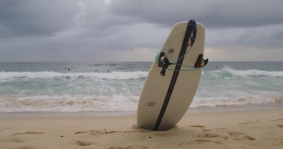 Surfboard stuck in the sand on a beach as the waves roll in under a cloudy sky