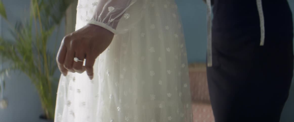 A bride with a diamond ring is shown getting her dress fitted by an assistant who has a tape measure