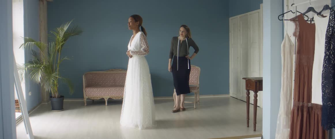 A woman in a wedding dress looks at her reflection in a mirror as a tailor watches her from behind