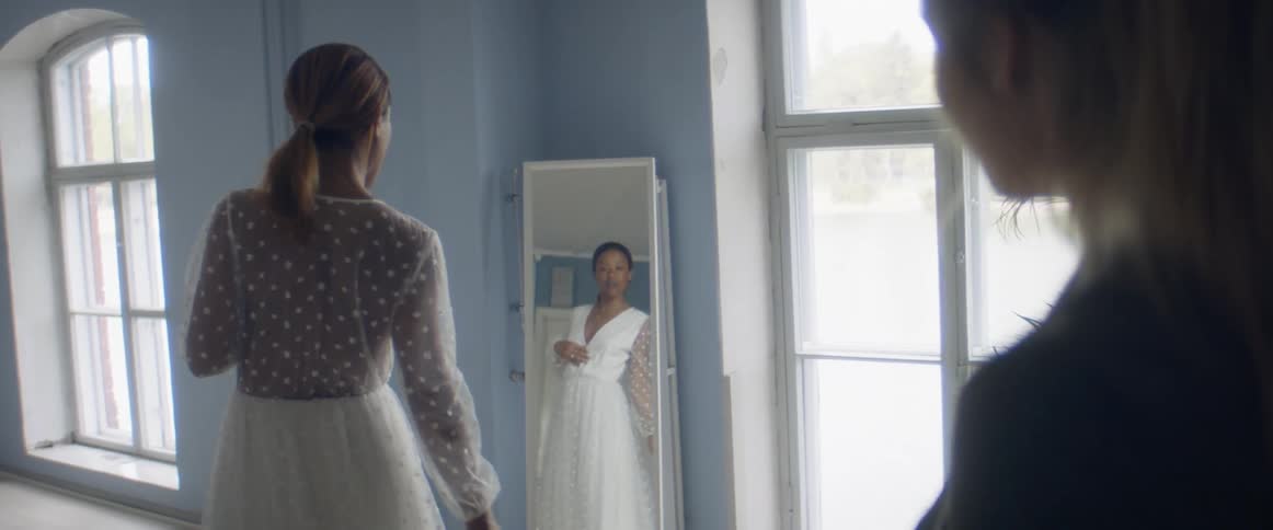 Two women in a room with one looking at a dress in a mirror with a wall in windows in the background