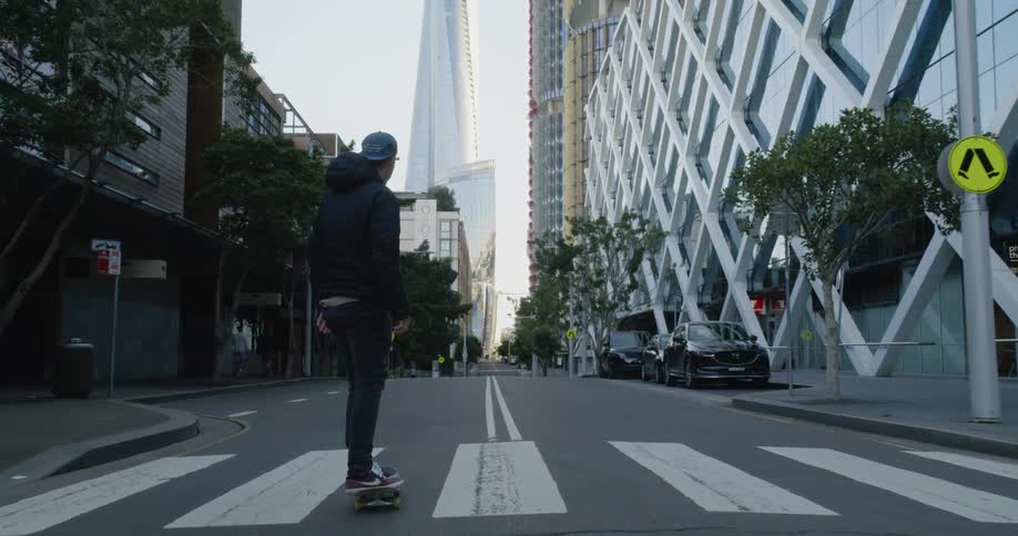 Man wearing jacket and baseball cap rides skateboard down empty city street past parked cars and skyscrapers