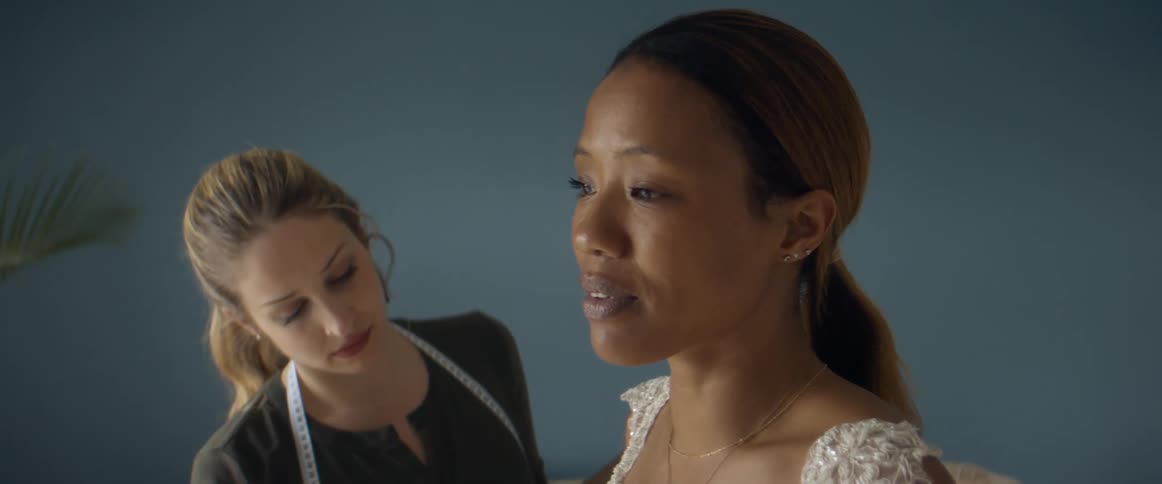 Two women in a room with one with a measuring tape and adjusting a dress on the other woman with a wall in the background