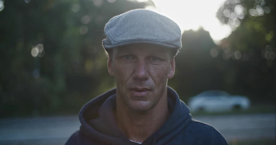 Man with tweed cap and blue hoodie faces the camera with eye contact next to highway