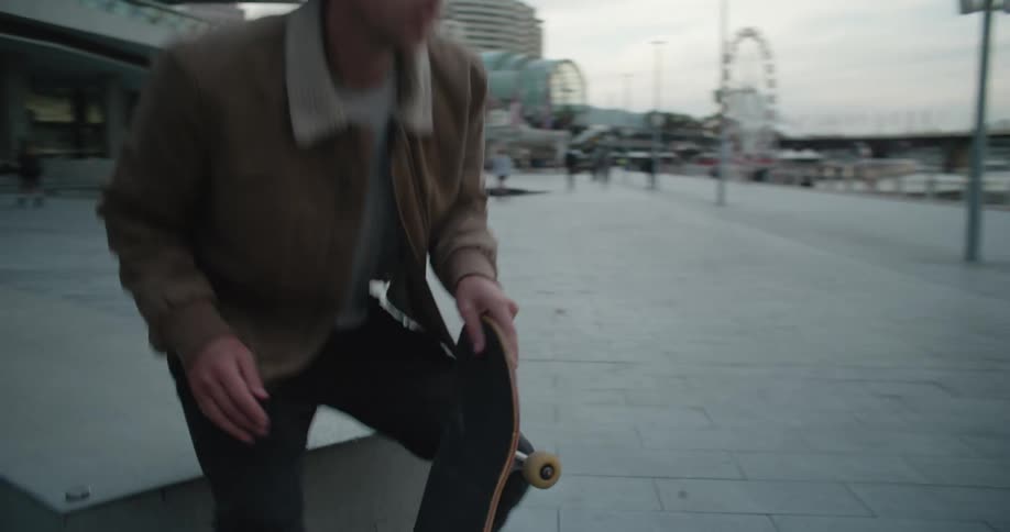 Man sitting looking over reflective water past marina and downtown skyscrapers stands to set off on skateboard