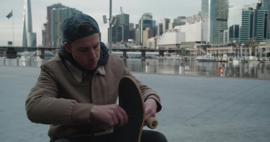 Man sitting near the harbour in the city adjusting the wheels on a skateboard