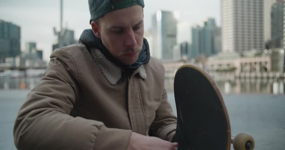 Man sitting near the harbour in the city adjusting the skateboard wheels