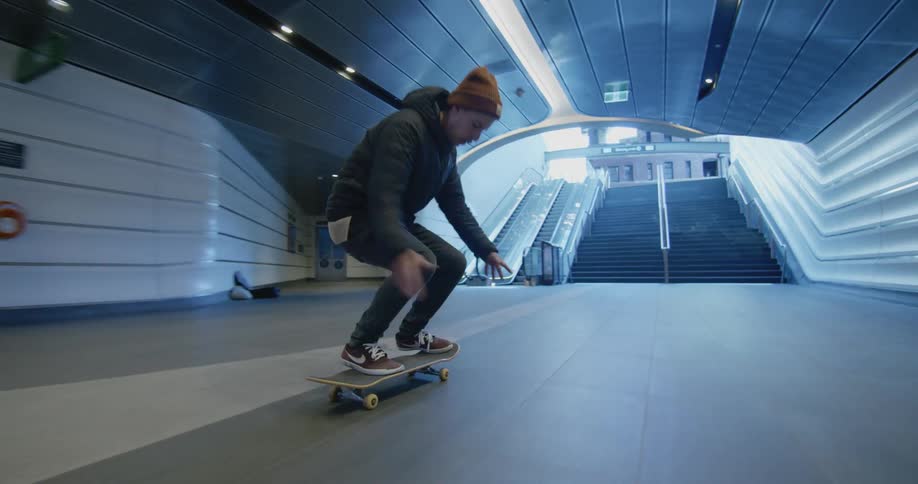 Man rides skateboard along wide passageway stopping at foot of stairs to ground level