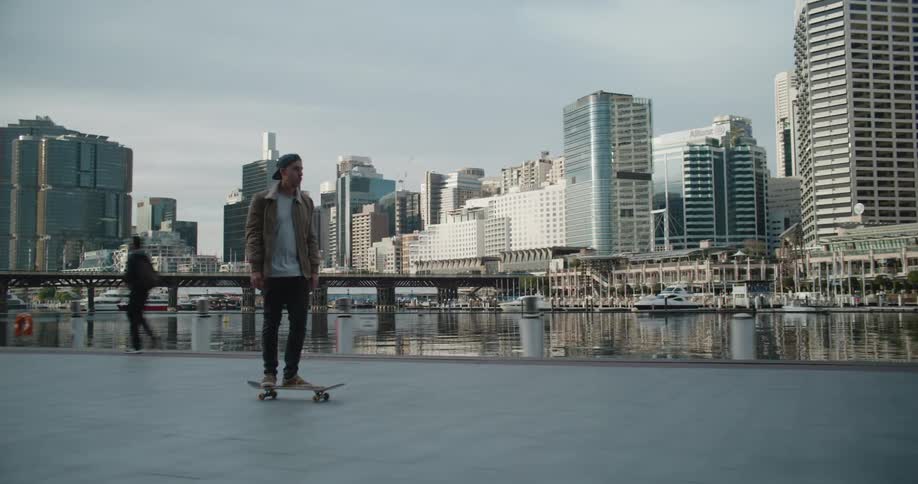 Skateboarder rides skateboard past pedestrians along walkway bordering river across from downtown skyscrapers