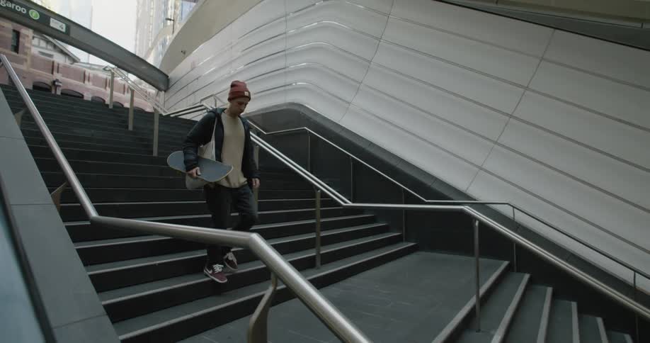 A man walking downstairs holding a skateboard with buildings the sky and clouds in the background