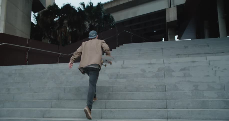 A man skating on a skateboard then picking it up and walking up stairs with an overpass the sky and clouds in the background