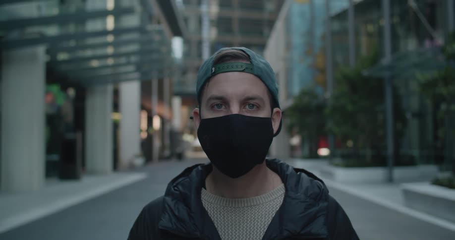 Man with a blue cap and black coat and mask looks at the camera in between city buildings