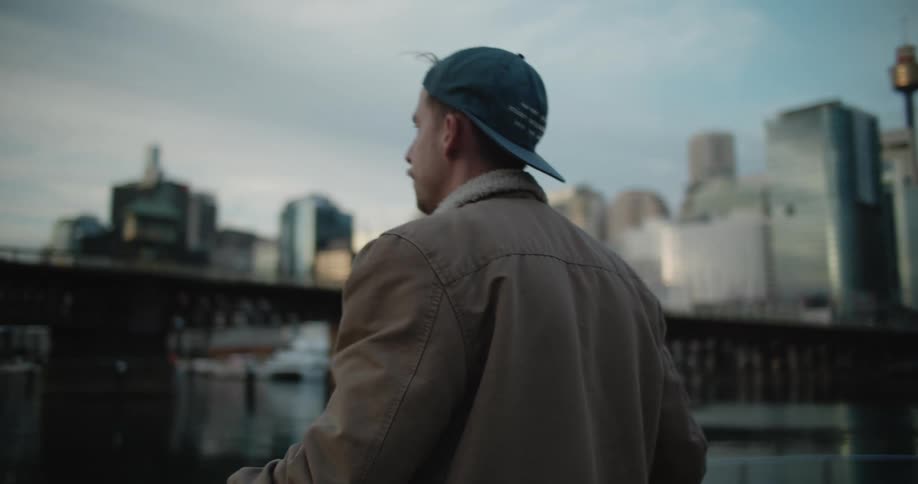 A man with a hat holding a skateboard near a dock with a bridge building the sky and clouds in the background