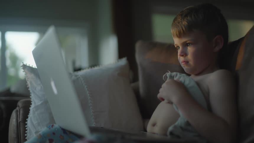 A boy is sitting on a couch and looking at a laptop