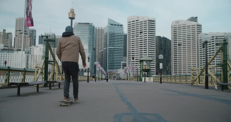 Man with a cap coat and grey pants skateboards on the bridge heading towards tall city buildings