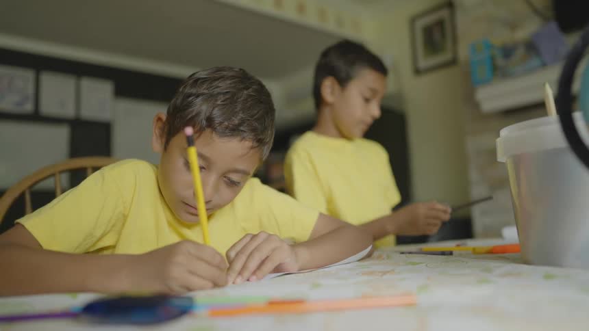Two male students with yellow shirts sitting at desk draw on papers in the classroom