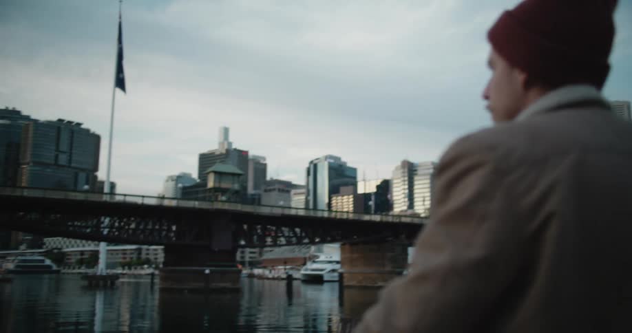 A man spins his skateboard in his hands while turning to look at a river in the city