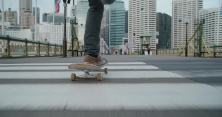 Man with brown shoes and jeans rides on skateboard with high rise buildings in the background