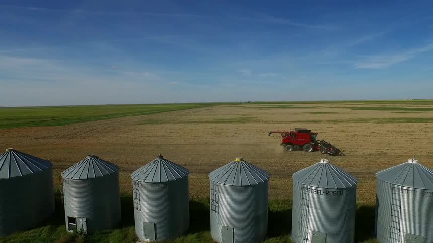 A row of grain silos stand before a field being worked by a tractor