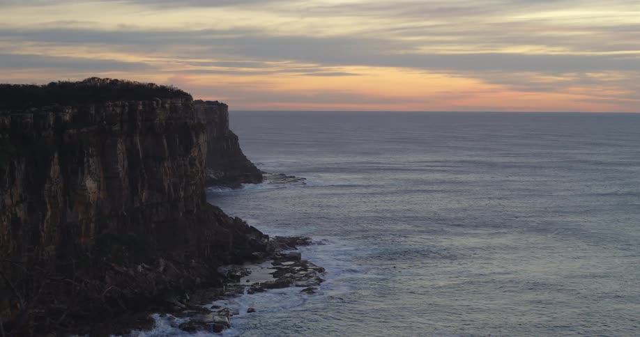 Waves crash against tall cliffs topped by forest against a pink tinted sky on cloudy day