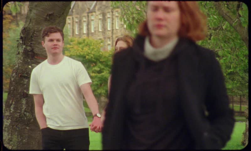 A young man and woman walk through a college campus holding hands as the people around them walk backward