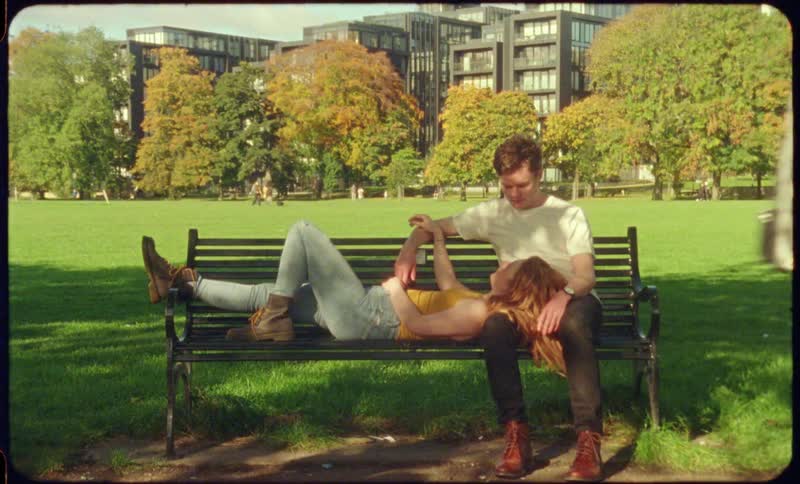A young woman lying on her boyfriend and chatting on a park bench as people walk by during the day