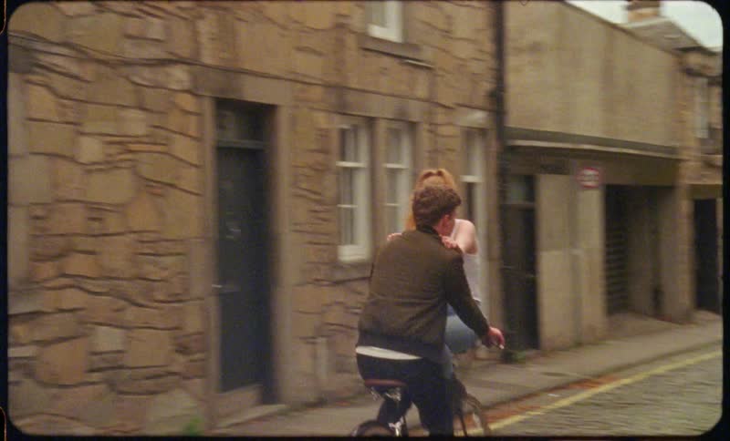Man pedals bicycle along narrow road between apartments as woman sits on handle bars