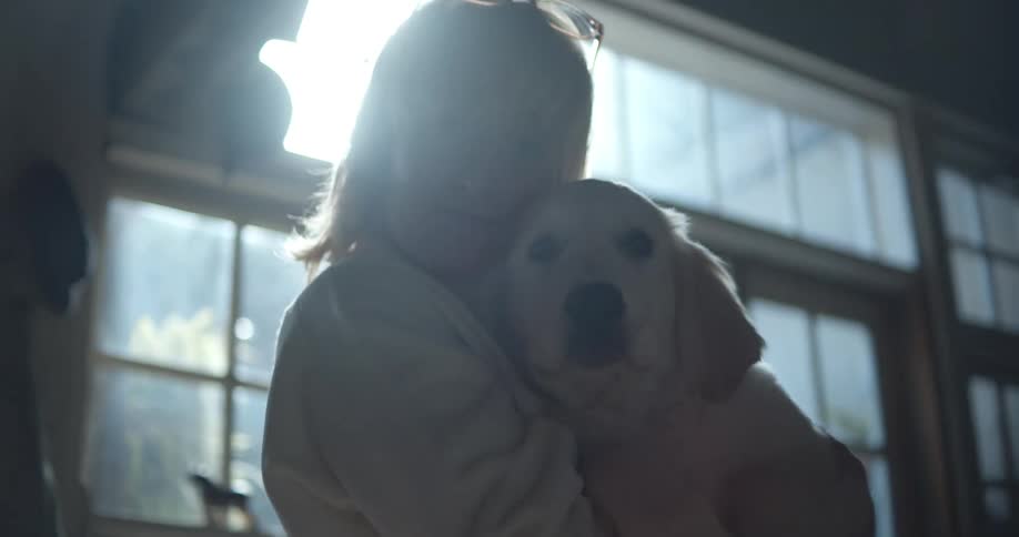 A woman with glasses holding a dog in a room with a wall and window in the background