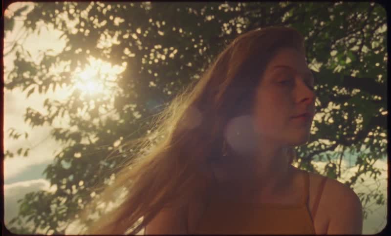 A woman sitting staring and smiling with a tree the sky and clouds in the background