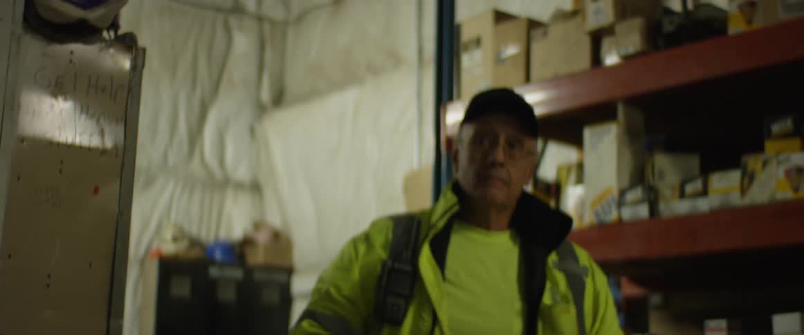 A man puts on a bright yellow reflective jacket with radio attached and walks away from work lockers