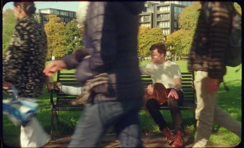 A young couple relax on a park bench as numerous people walk past