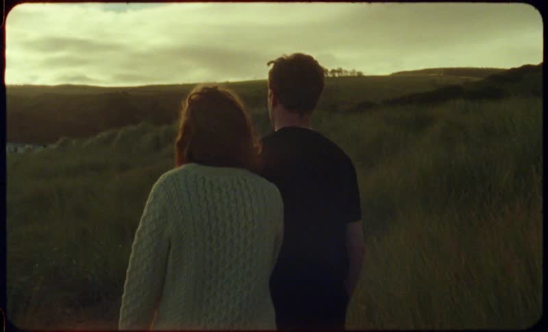 A man and woman hold hands while walking through a field together in the evening