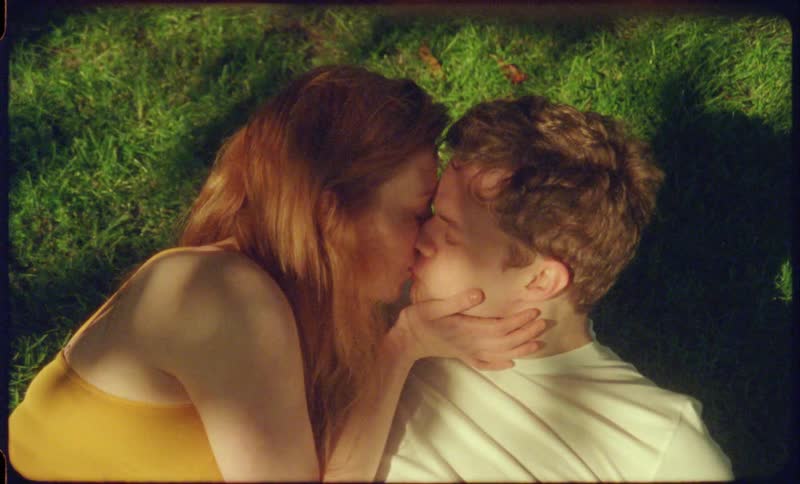 A young woman leans over to kiss her male companion who is relaxing in the shade of a tree