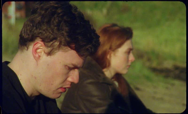 Woman sits near a hill looking angry while a man sits next to her looking sad and remorseful