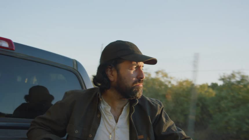 A man with a hat riding in the back of a truck with trees the sky and clouds in the background
