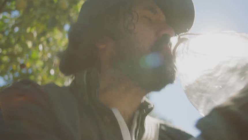 Bearded man drinks water from a jug while standing near the forest under the bright sun