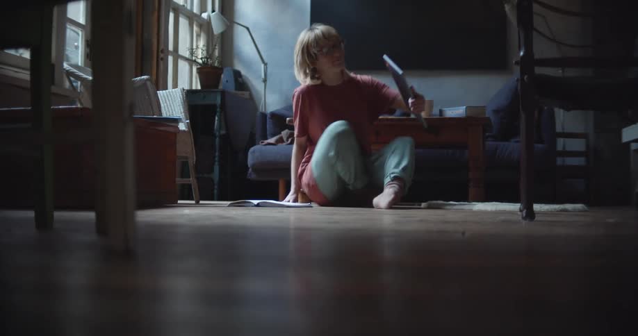 Woman walks across the floor sets a bowl on a table then sits on the floor with a laptop as a dog rushes over to her