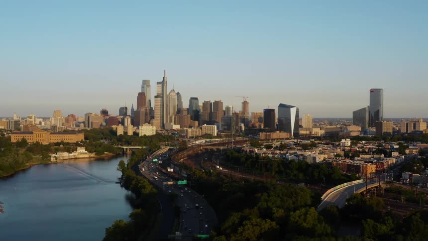 Traffic moves along highway paralleling reflective river leading to downtown Philadelphia skyscrapers