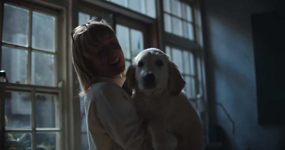 A woman with glasses holding a dog in a room with a wall and window in the background