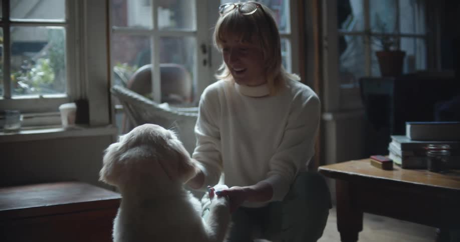 A woman feeds and pets a dog while kneeling in front of it in the living room of a house during the day