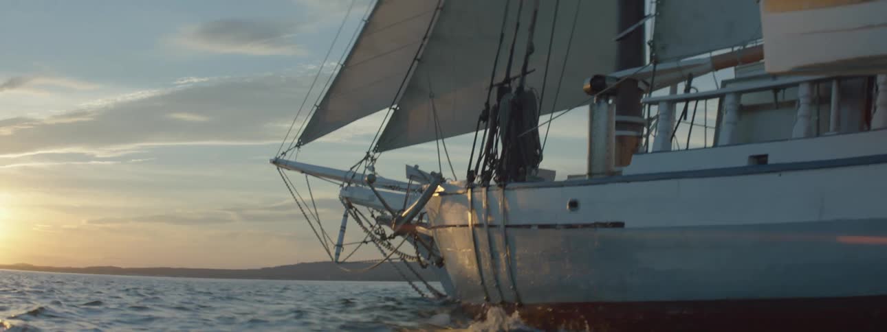 Sun hovers on misty horizon over ocean as boat with sails unfurled sails through ocean