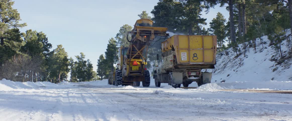 A tractor and a dump truck clearing snow on a road between a surrounding forest during the day