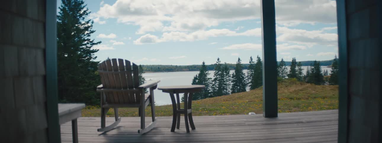A chair and table sit on the front porch of a cabin on a lake