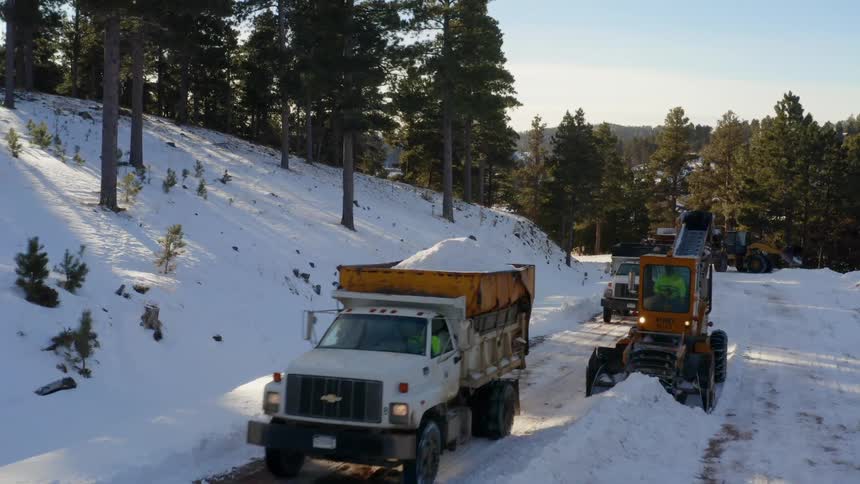 Dump trucks drive along snowy road past snow loading machine near slope and forest