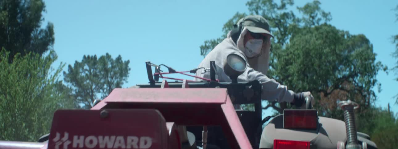 A person with a mask driving a tractor with trees the sky and clouds in the background