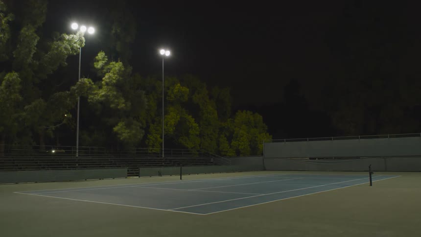 A tennis court with light posts trees and bleachers with vehicles and the sky in the background