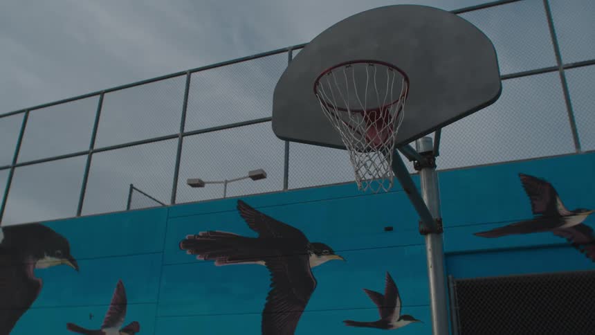 A basketball hoop on an empty court with a mural painted wall and fence behind