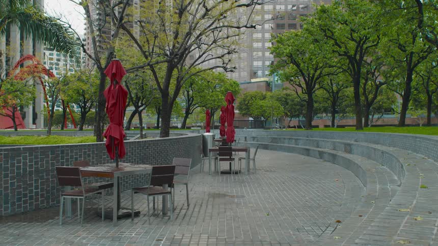 Tables with folded red umbrellas stand in park surrounded by buildings