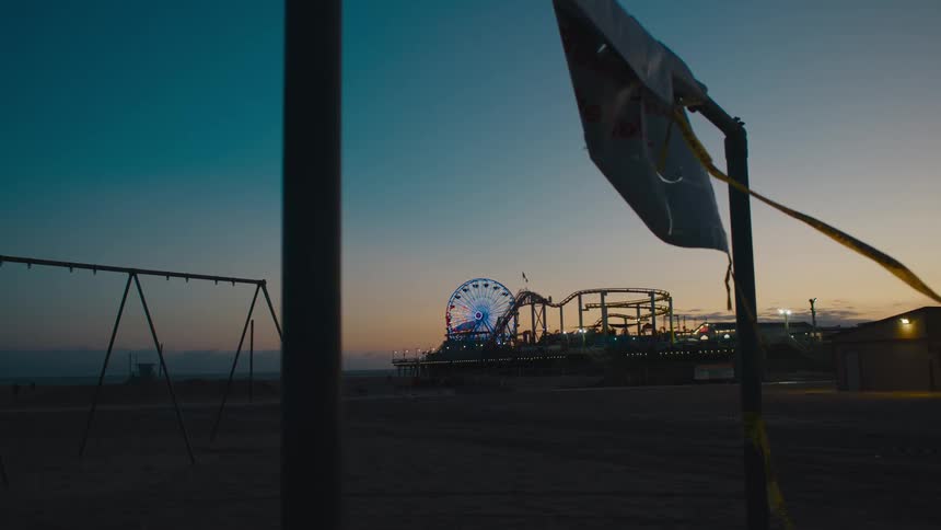 Empty swing set frame stands under red tinted sky at night as list ferris wheel and roller coaster set in background