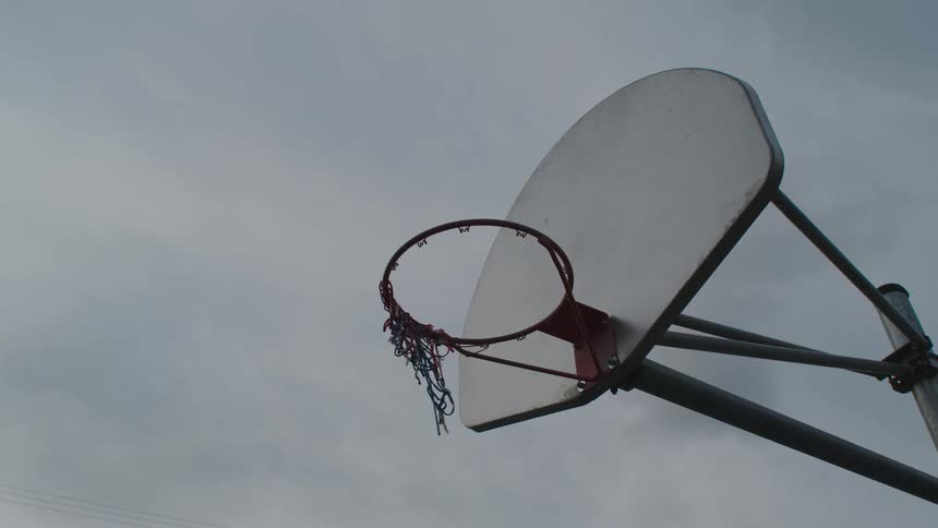 Remains of torn netting flutters in breeze on basketball hoop under overcast sky